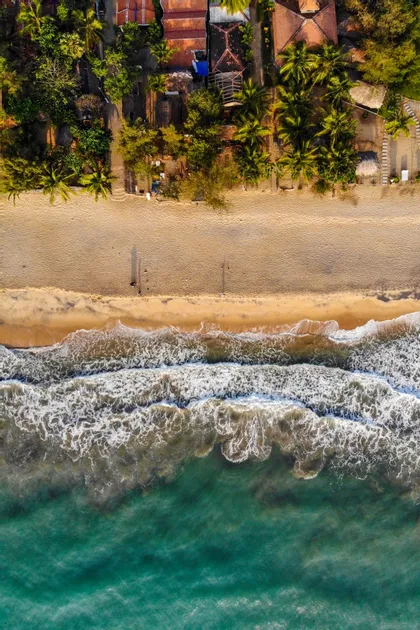 Veduta aerea dall'alto di onde turchesi che lambiscono una spiaggia sabbiosa, bordata da palme ed edifici.