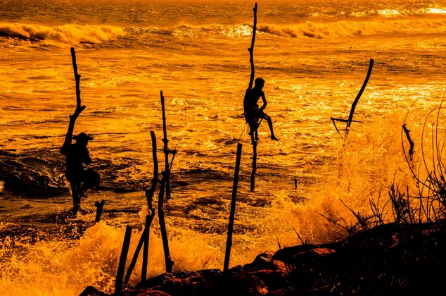 Sagome di due pescatori su palafitte in un mare mosso, arancione dorato, al tramonto, con le onde che si infrangono.