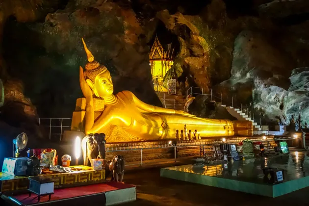 Une grande statue de Bouddha couché doré, illuminée par des lumières artificielles à l'intérieur d'une grotte sombre et rocheuse.