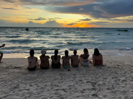 Un groupe WeRoad de femmes assises sur une plage, face à l'océan, regardant le coucher du soleil.