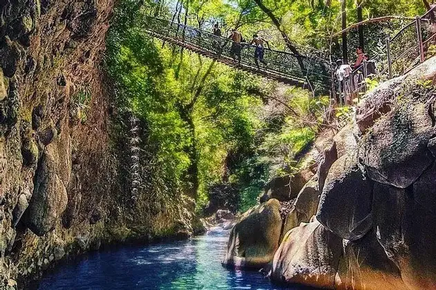 Un viaggio di gruppo WeRoad attraversa un ponte sospeso su un fiume turchese che scorre attraverso un canyon roccioso e alberato.