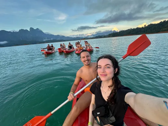 Un groupe WeRoad prend un selfie en kayak dans des bateaux rouges sur un lac, avec des montagnes en arrière-plan sous un ciel nuageux.