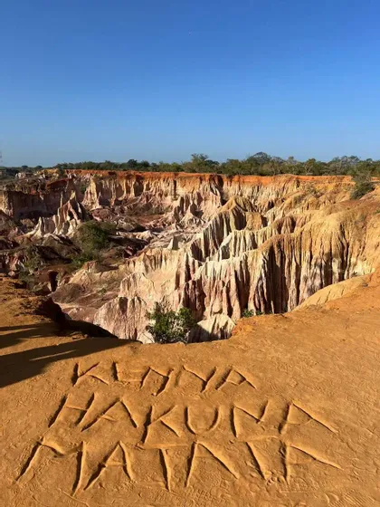 Le parole 'Kenya Hakuna Matata' incise nella sabbia arancione sul bordo di una scogliera, con vista su un vasto canyon multicolore.