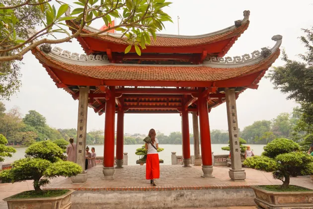 Una donna in pantaloni rossi si trova all'interno di una tradizionale pagoda rossa con tetto a tegole, guardando fuori su un lago con alberi e bonsai.