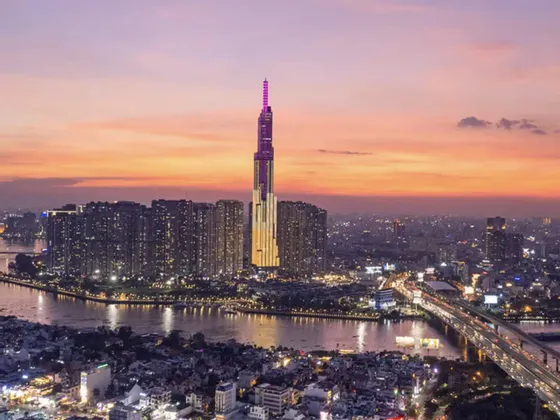 Un grattacielo illuminato con una guglia viola domina un denso skyline cittadino accanto a un ampio fiume al tramonto.