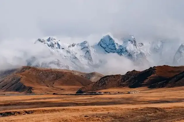 Des pics montagneux enneigés percent les nuages au-dessus d'une vaste steppe dorée, avec un petit village au loin.