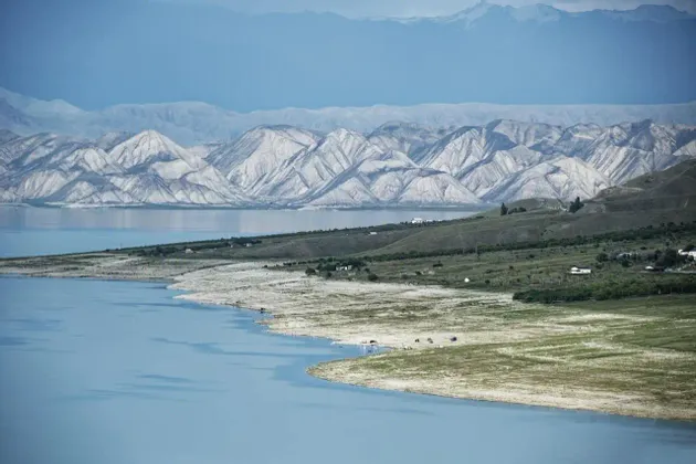 Un vasto lago azul se curva a lo largo de una orilla de verdes colinas, con capas de cordilleras blancas y azules al fondo.