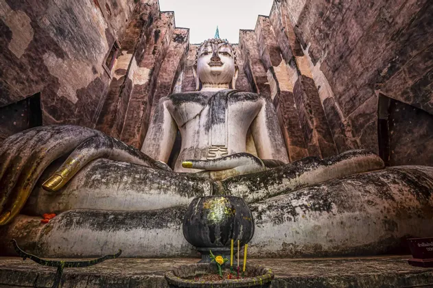 Eine kolossale, verwitterte Stein-Buddha-Statue sitzt meditierend zwischen hohen Steinmauern, mit einer kleinen Opferschale davor.
