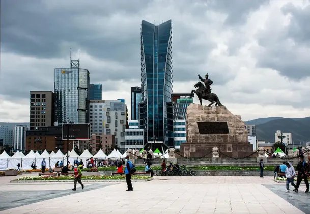 Eine Reiterstatue auf einem großen Steinsockel auf einem Stadtplatz, mit modernen Wolkenkratzern im Hintergrund unter einem bewölkten Himmel.