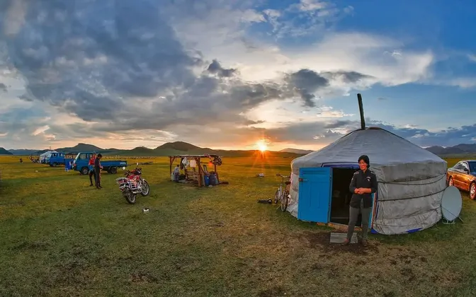 Bei Sonnenuntergang steht eine Person vor einer traditionellen Jurte auf einem Campingplatz auf einer grasbewachsenen Ebene, im Hintergrund Fahrzeuge und Berge.