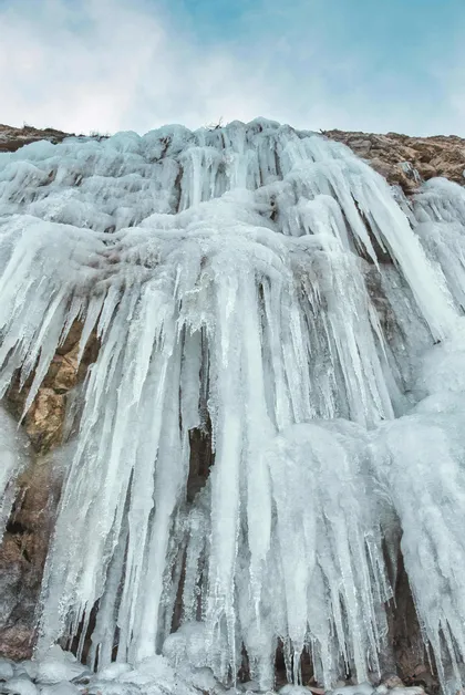 Eine Tiefenansicht eines gefrorenen Wasserfalls mit zahlreichen Eiszapfen, die an einer felsigen Klippenwand unter einem bewölkten Himmel hängen.