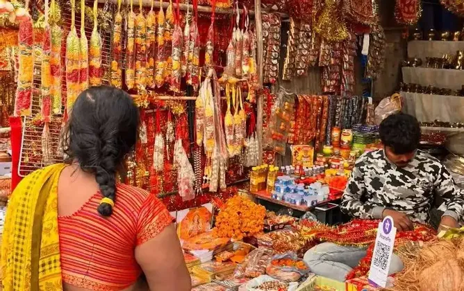 Una donna in sari osserva una bancarella in un mercato all'aperto, piena di ghirlande colorate e articoli religiosi venduti da un uomo.