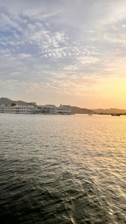Un grande edificio bianco affacciato su un lago al tramonto, con colline lontane sotto un cielo parzialmente nuvoloso.