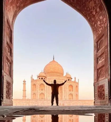 Una persona in silhouette a braccia alzate si erge davanti al Taj Mahal, visto attraverso un arco ornato con un riflesso in primo piano.