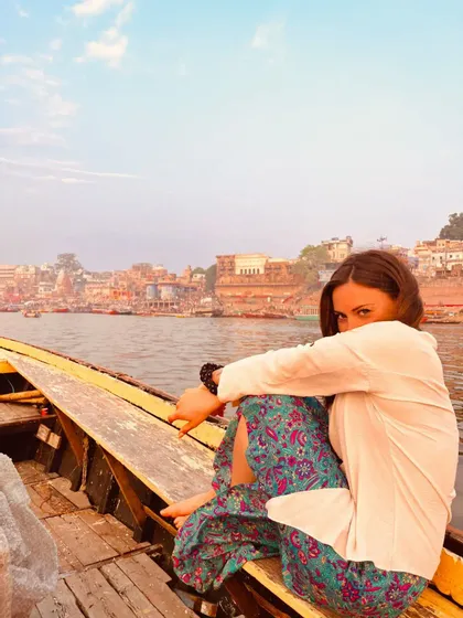 Una donna con pantaloni a motivi colorati siede in una barca di legno su un fiume, con uno skyline cittadino sullo sfondo.