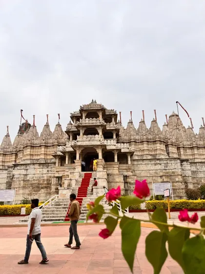 Un tempio in pietra ornato con una scalinata rivestita di tappeto rosso, con persone nel cortile e fiori rosa in primo piano.