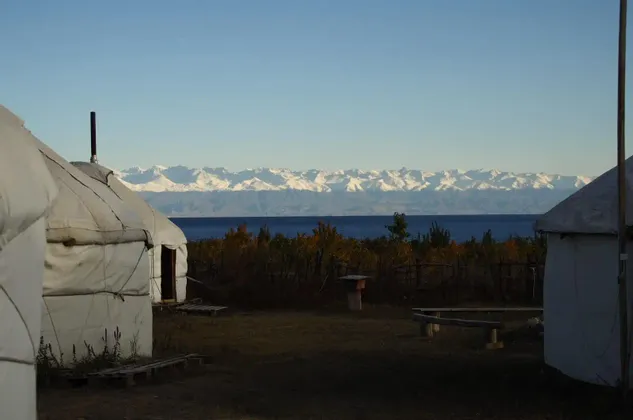 Yurtas blancas a la orilla de un lago con una cordillera nevada en la distancia bajo un cielo despejado.