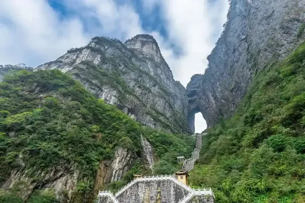 Una larga escalera asciende por una montaña empinada y cubierta de bosque hacia un gran arco de roca natural bajo un cielo nublado.