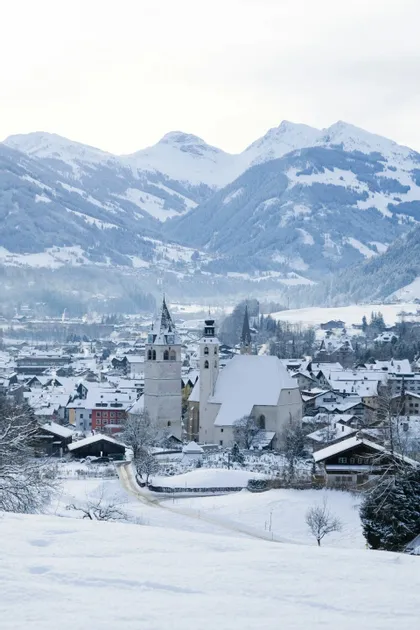Blick von oben auf ein schneebedecktes Alpendorf mit einer markanten Kirche vor schneebedeckten Bergen.