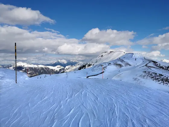 Ein schneebedeckter Berghang mit Skispuren im Vordergrund und entfernten Skifahrern unter blauem Himmel mit weißen Wolken.