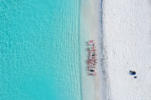 Vista aérea de un grupo de WeRoad tumbado en fila en una playa de arena blanca junto a aguas turquesas y cristalinas.