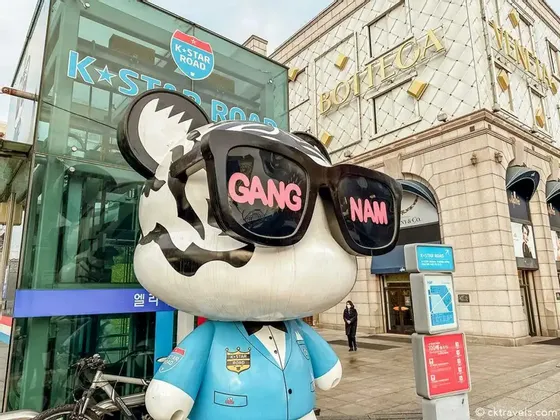 Una grande statua di orso che indossa una giacca blu e occhiali da sole con su scritto 'GANG NAM' si trova su una strada cittadina davanti all'insegna della K-Star Road.