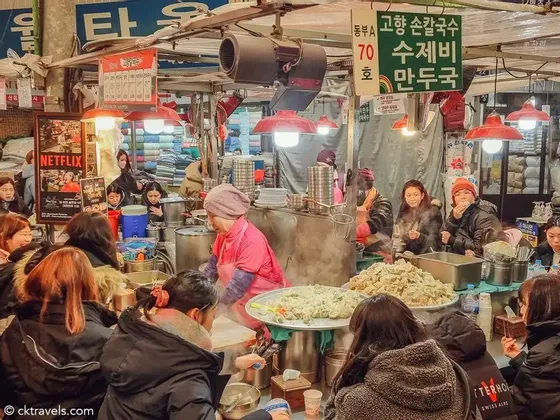 Un viaggio di gruppo WeRoad che mangia a una bancarella di un affollato mercato alimentare coperto mentre un venditore prepara ravioli fumanti.