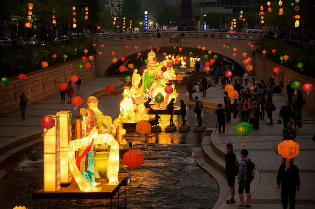 Una grande lanterna illuminata galleggia su un fiume di notte durante un festival, con persone che camminano sulle rive.