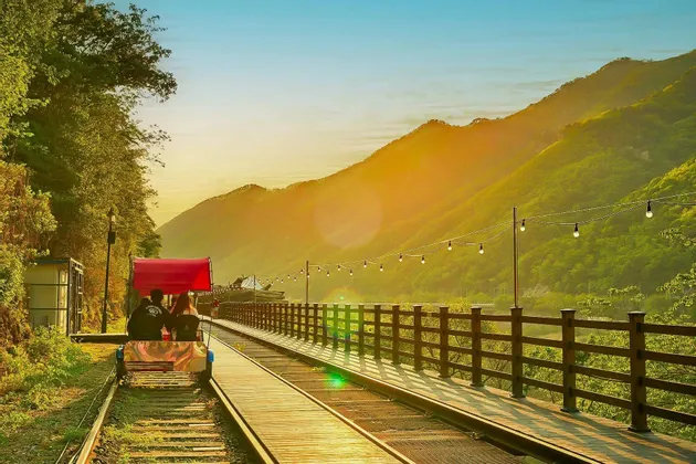 Una coppia percorre una bici su rotaia con una tettoia rossa lungo un binario attraverso una valle di montagna durante un tramonto dorato.