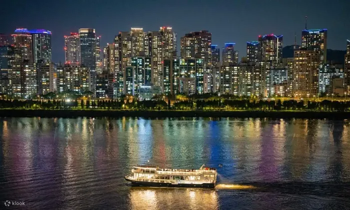 Una barca illuminata naviga di notte su un fiume, di fronte a uno skyline cittadino illuminato.