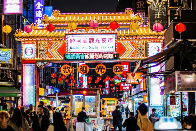 Un arco tradizionale illuminato con lanterne appese segna l'ingresso di un vivace mercato notturno cittadino, affollato di persone.