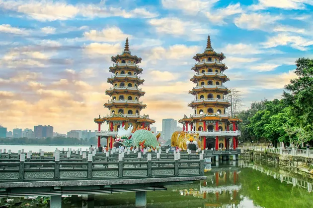 Due pagode ornate con ingressi di drago e tigre si ergono su un lago, con uno skyline cittadino e un cielo nuvoloso sullo sfondo.