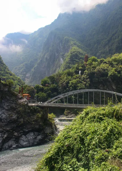 Un ponte ad arco in acciaio attraversa una gola fluviale rocciosa, circondato da montagne verdi e lussureggianti, con una pagoda visibile in lontananza.
