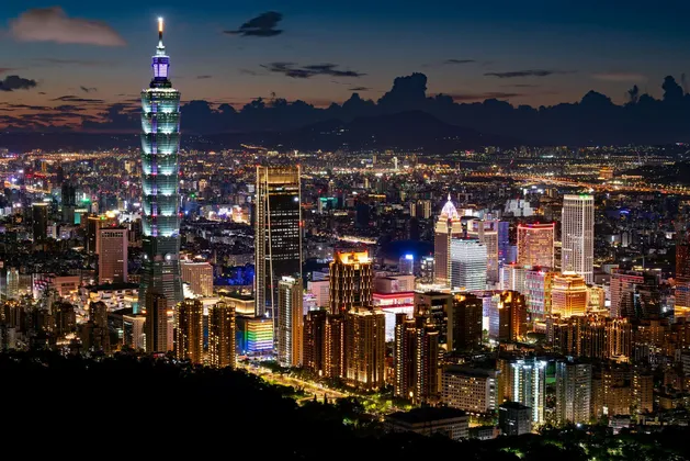 Una vista panoramica di uno skyline cittadino esteso e illuminato di notte, con un alto e distintivo grattacielo che si erge sopra gli altri edifici.