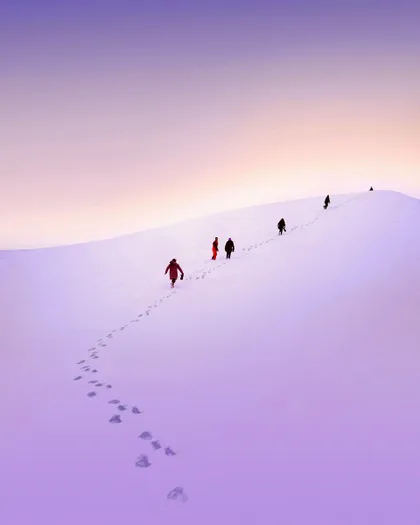 Un viaggio di gruppo WeRoad cammina in fila indiana su un vasto pendio innevato, lasciando impronte sotto un cielo viola e arancione.