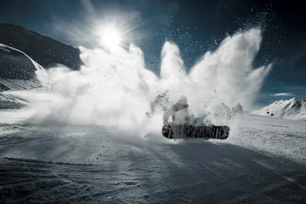 Uno snowboarder traccia la neve fresca su un pendio di montagna, sollevando un'enorme nuvola di neve nell'aria sotto un sole splendente.