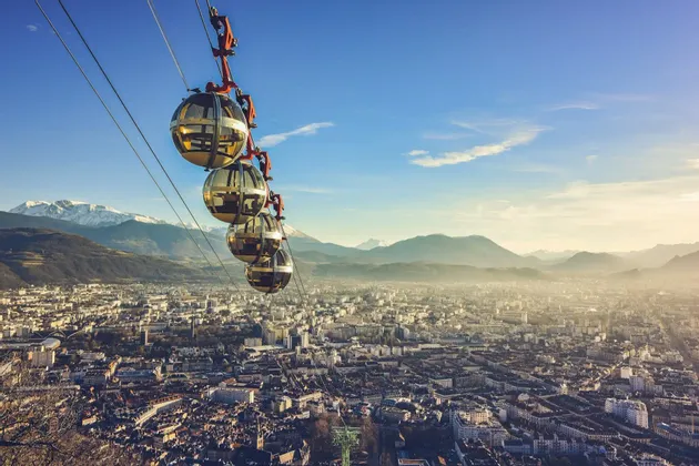 Una fila di funivie sferiche viaggia sopra una città estesa, con uno sfondo di montagne sfocate e cime innevate in lontananza.