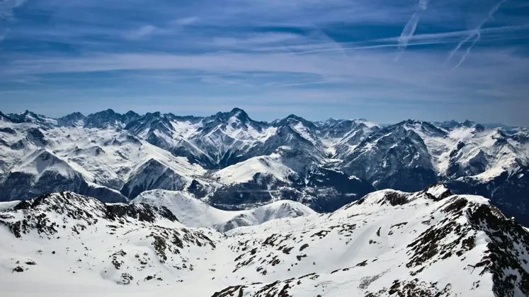 Una vista panoramica di una vasta catena montuosa innevata sotto un cielo azzurro con nuvole sottili e scie di condensazione.