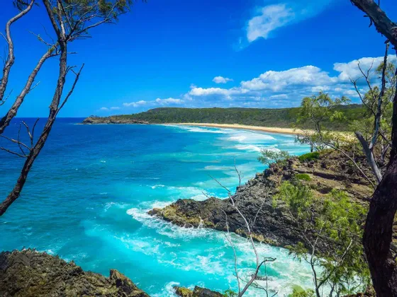 Ein Blick von einem hohen Aussichtspunkt auf eine tropische Küste mit türkisfarbenem Wasser, einem Sandstrand und grünen Hügeln.