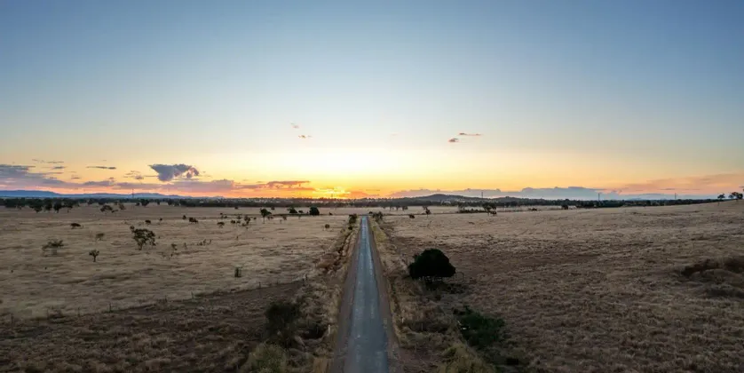 Luftaufnahme einer geraden Straße, die sich durch eine weite, trockene Savanne dem Sonnenuntergang am Horizont entgegenzieht.