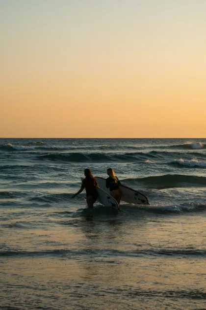Due donne con tavole da surf camminano tra le onde dell'oceano sotto un caldo cielo al tramonto.