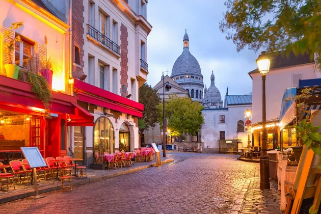 Una strada acciottolata con caffè dalle luci calde e una grande basilica dalla cupola bianca sullo sfondo al tramonto.