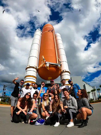 Eine WeRoad-Gruppe posiert für ein Foto vor der Nachbildung einer Weltraumrakete unter bewölktem Himmel.