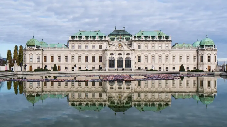 Un grande palazzo bianco e sontuoso con un tetto verde si riflette in uno specchio d'acqua immobile in primo piano.