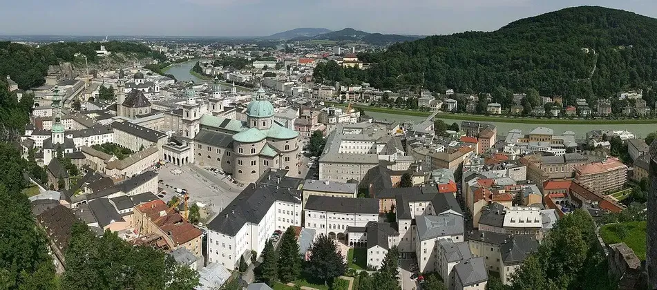 Una vista panoramica dall'alto di una storica città europea con una grande cattedrale, un fiume e una collina boscosa sullo sfondo.