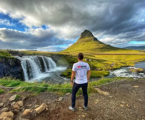 Un capogruppo WeRoad visto di spalle, che osserva una cascata che scorre accanto a una grande montagna verde sotto un cielo nuvoloso.