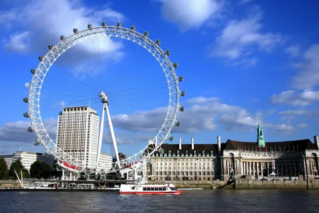 Una grande ruota panoramica bianca si erge sulla riva di un fiume accanto agli edifici della città, con una barca rossa e bianca sull'acqua.