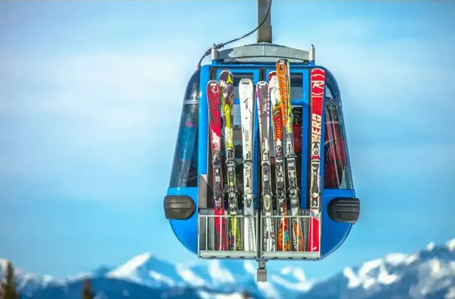Eine blaue Skigondel mit Skiern im Außenfach fährt unter klarem blauem Himmel über eine verschneite Bergkette.