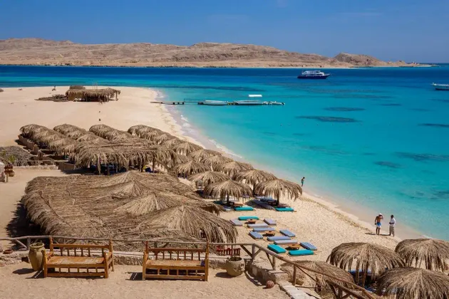 Vue plongeante sur une plage tropicale, avec des parasols en paille le long du rivage aux eaux turquoise cristallines et des collines désertiques en arrière-plan.