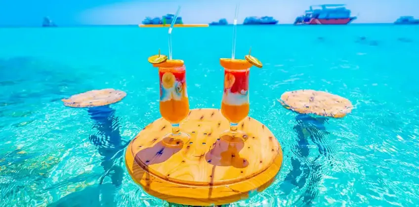 Deux cocktails aux fruits sur une petite table en bois flottant dans une eau turquoise claire, avec des bateaux visibles à l'horizon sous un ciel lumineux.